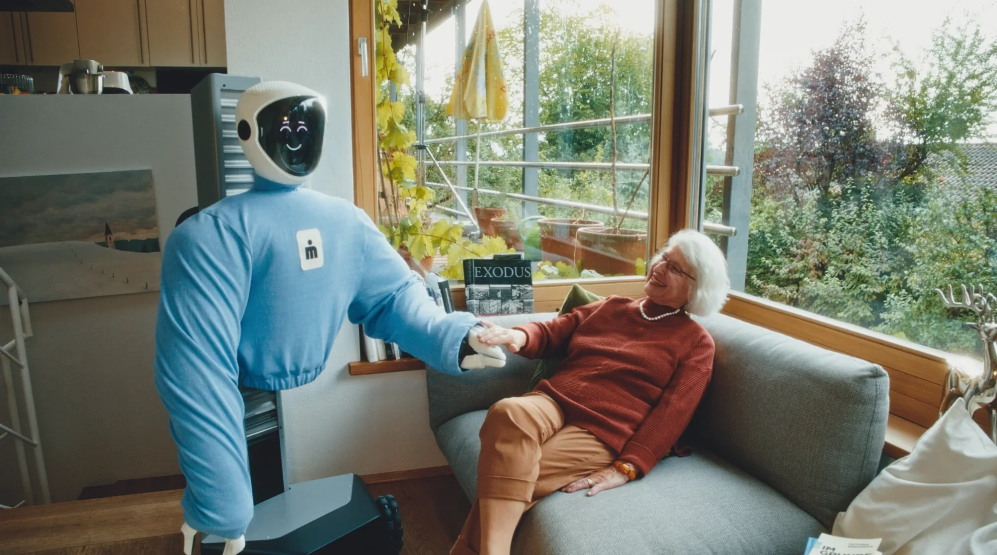 Plim reaching out to hold the hand of an older woman on her sofa.